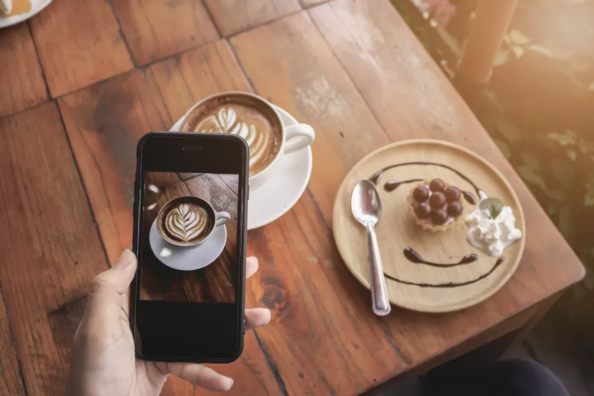 Ahşap masada latte art süslemeli kahvenin ve tatlının fotoğrafını akıllı telefonuyla çeken bir kişinin elleri