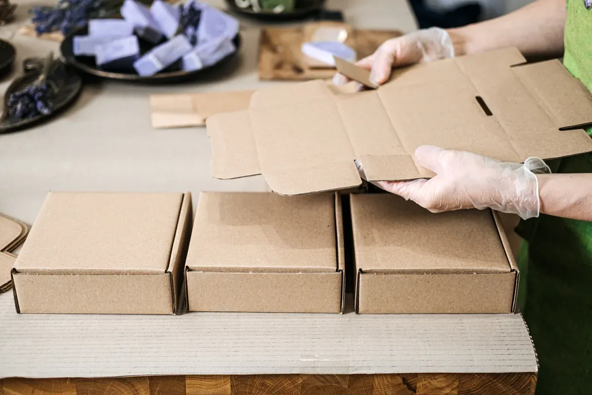 Eldivenli ellerle masada kraft karton kutuları katlayıp online siparişleri paketlemeye hazırlayan bir kişi