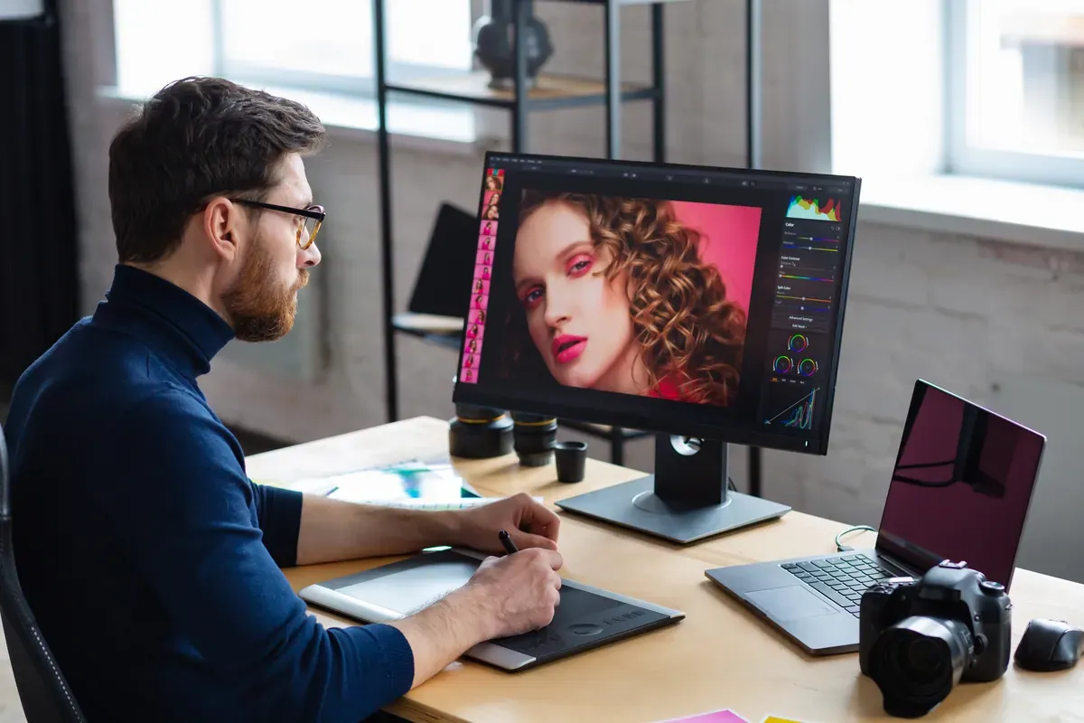 Ofisinde monitör, dizüstü bilgisayar ve grafik tabletle bir portre fotoğrafına rötuş yapan gözlüklü grafik tasarımcı