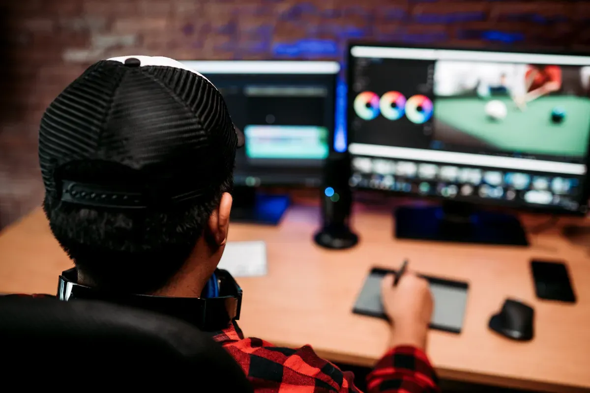 Ev stüdyosunda şapkalı bir video editörünün arkadan görünümü ve monitörlerde açık duran kurgu ve renk ekranları