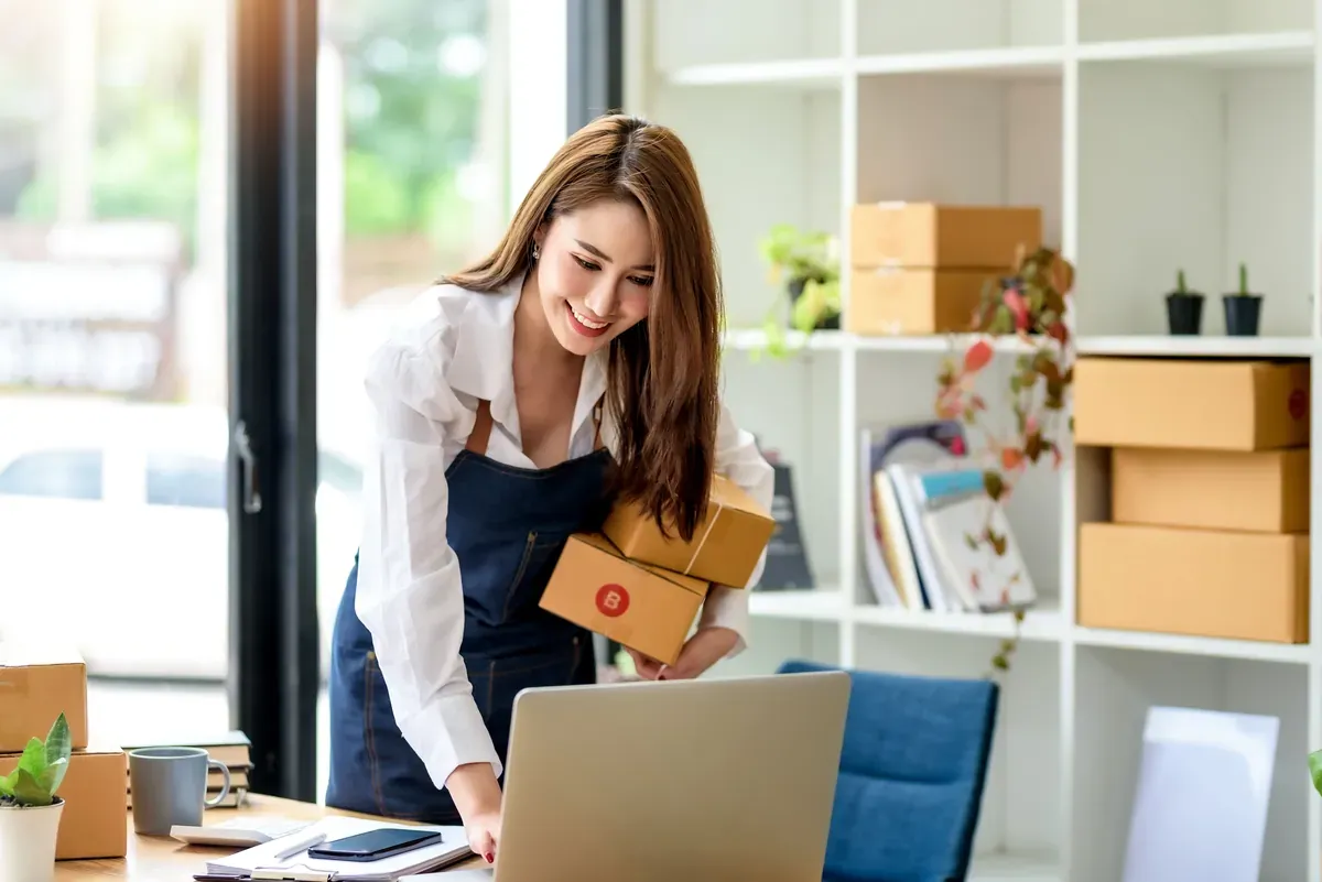 Aydınlık çalışma odasında karton koli hazırlarken dizüstü bilgisayardan online siparişleri kontrol eden genç kadın